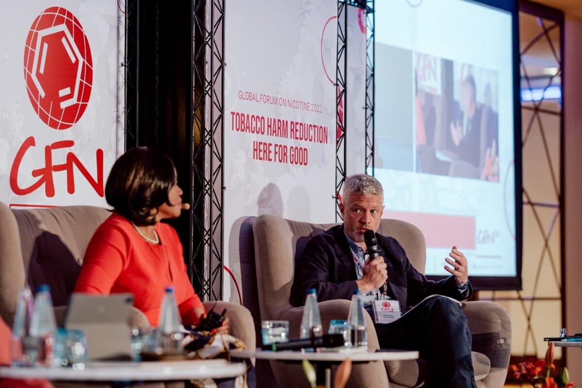 Two people sit on a stage and lead a discussion during a conference. The man is speaking into a microphone and the woman is listening intently. In the background are the captions "Global Forum on NICOTINE 2023" and "Tobacco Harm Reduction Here for Good" with the red GFN logo visible, making it perfect for a photo essay of the event.  