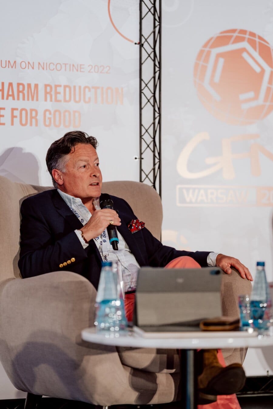 A man in a jacket, light-colored shirt and red pants sits in a chair and holds a microphone. Behind him is a background with text about nicotine harm reduction and the "GFN Warsaw 2022" logo. On the table in front of him is a laptop and bottles of water. The event was captured by a photographer at the conference.   