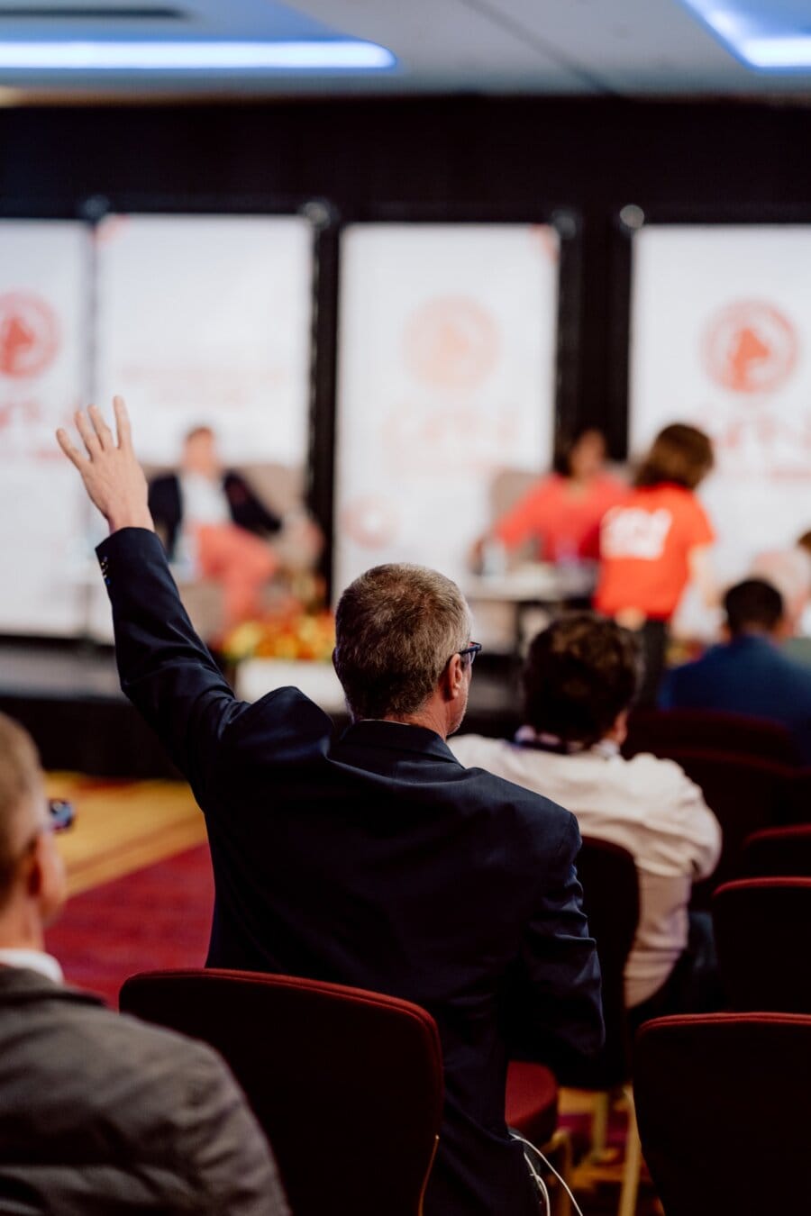 A person in a dark suit raises his hand from above the audience during a conference or panel discussion. Behind them on the stage, several speakers in red T-shirts can be seen with a fuzzy banner in the background. The scenery seems formal, reflecting an authentic photo-recording of the conference.  