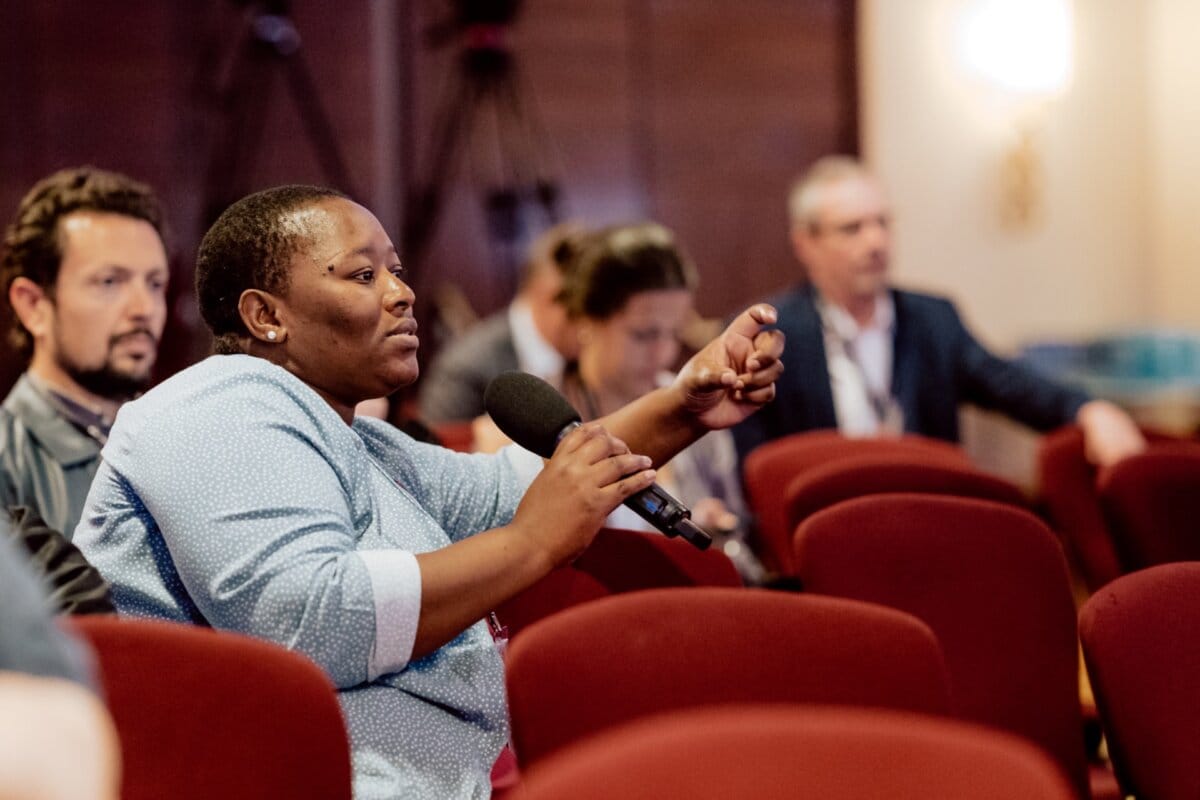 A person in a blue shirt speaks into a microphone during a conference or meeting. They sit among other attendees, who are also involved, in a room with red chairs and a fuzzy background, capturing moments for a photo essay of the event. 