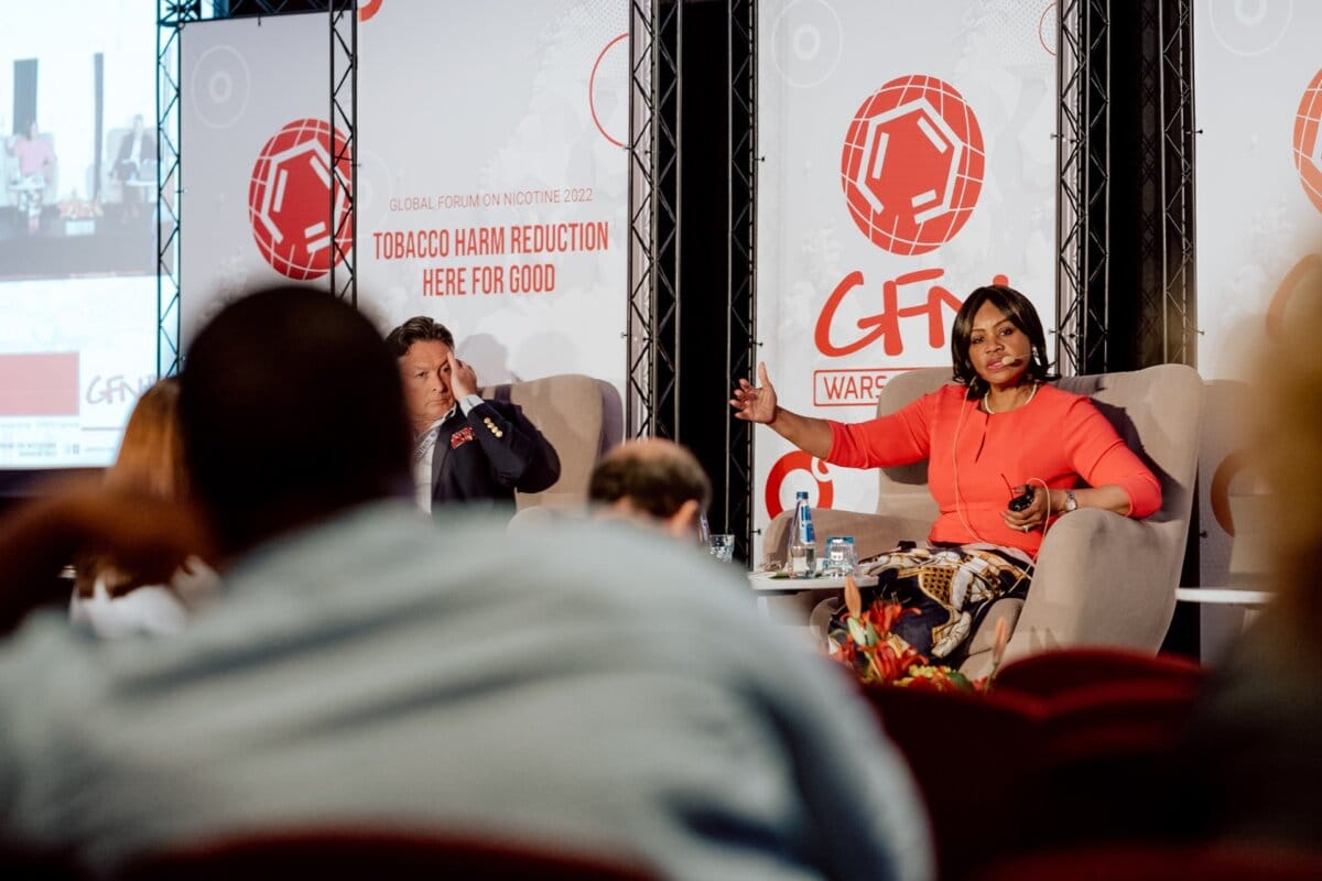 A woman in a red dress speaks as she sits on stage next to a man in a suit at the World Nicotine Forum 2022. Behind them hang banners reading "We will reduce the harms of smoking for good." Onlookers can be seen in the foreground, capturing a moment of extensive coverage of the event.  