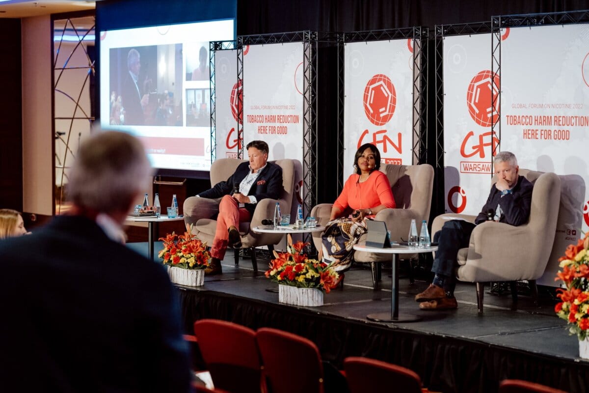 During the conference, a three-person panel sits on stage, as beautifully captured by a photo report of the event. Two men and one woman sit in chairs behind a table with bottles of water. The conference name and logo are displayed in the background, while a member of the audience is blurred in the foreground and the presentation screen is visible.  