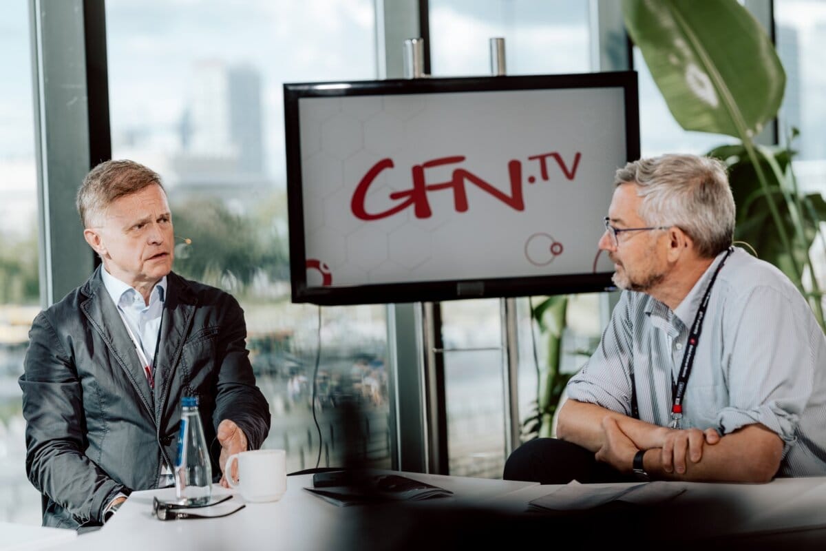 Two people are sitting at a table, with a "GFN.TV" sign in the background discussing. They look like they are in a studio, with large windows and greenery visible outside. The person on the left is gesturing with one hand. This scene could easily be part of a photo essay of the conference.   