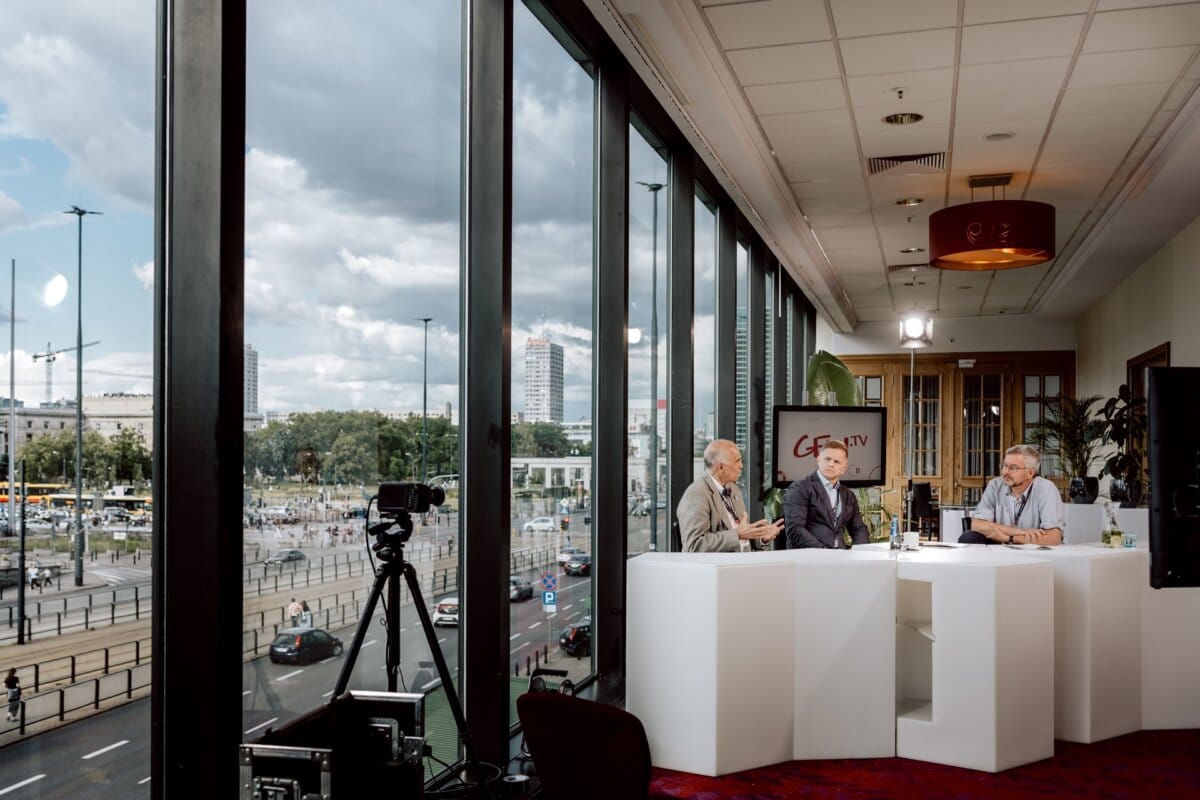 Three men in suits sit at a white table in a modern office with large floor-to-ceiling windows. They appear to be participating in a discussion or interview with recording equipment visible. Outside the windows is a vibrant cityscape that captures the dynamic atmosphere of the conference photo essay.  