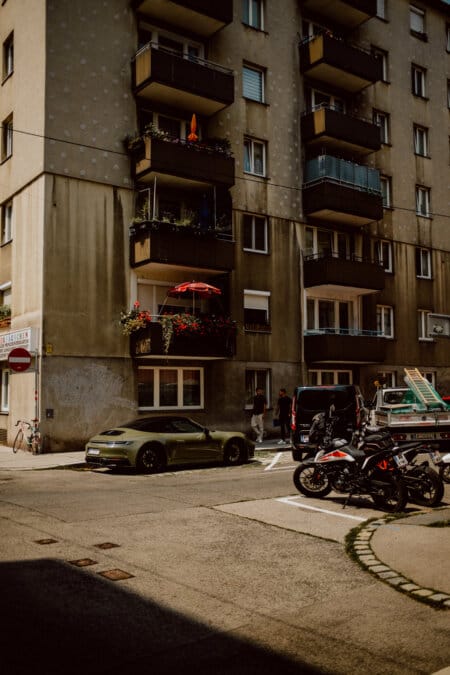 A street scene of a city with a multi-story apartment building with balconies decorated with plants and a red umbrella. Various vehicles are parked in the foreground, including motorcycles, a bicycle, a green sports car and a black van. The weather is sunny.  