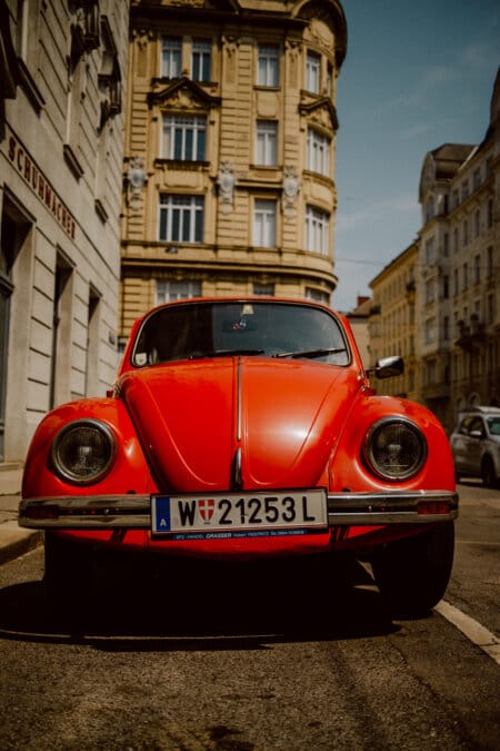 A red vintage Volkswagen Beetle parked on a street surrounded by historic European buildings. The car has a front license plate with the code "W 21253L" and a small flag symbol. On this bright day, the scene is illuminated by sunlight.  