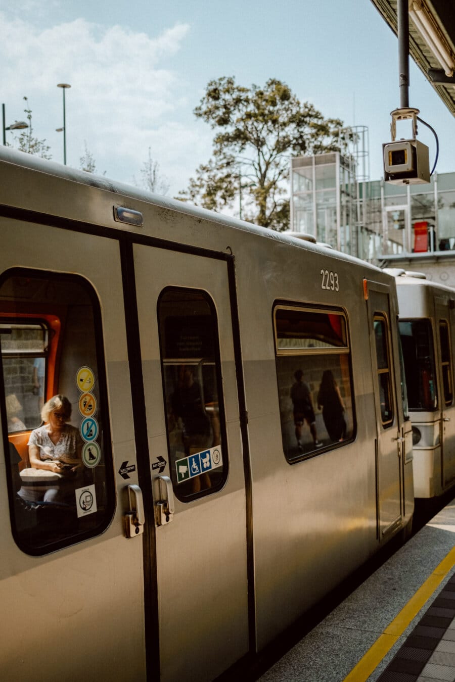 The train stops on the platform under a partly cloudy sky. A person sits on the train by the window while others walk on the platform outside. The train's door displays various signs, including priority seating designations. Trees can be seen in the background.   