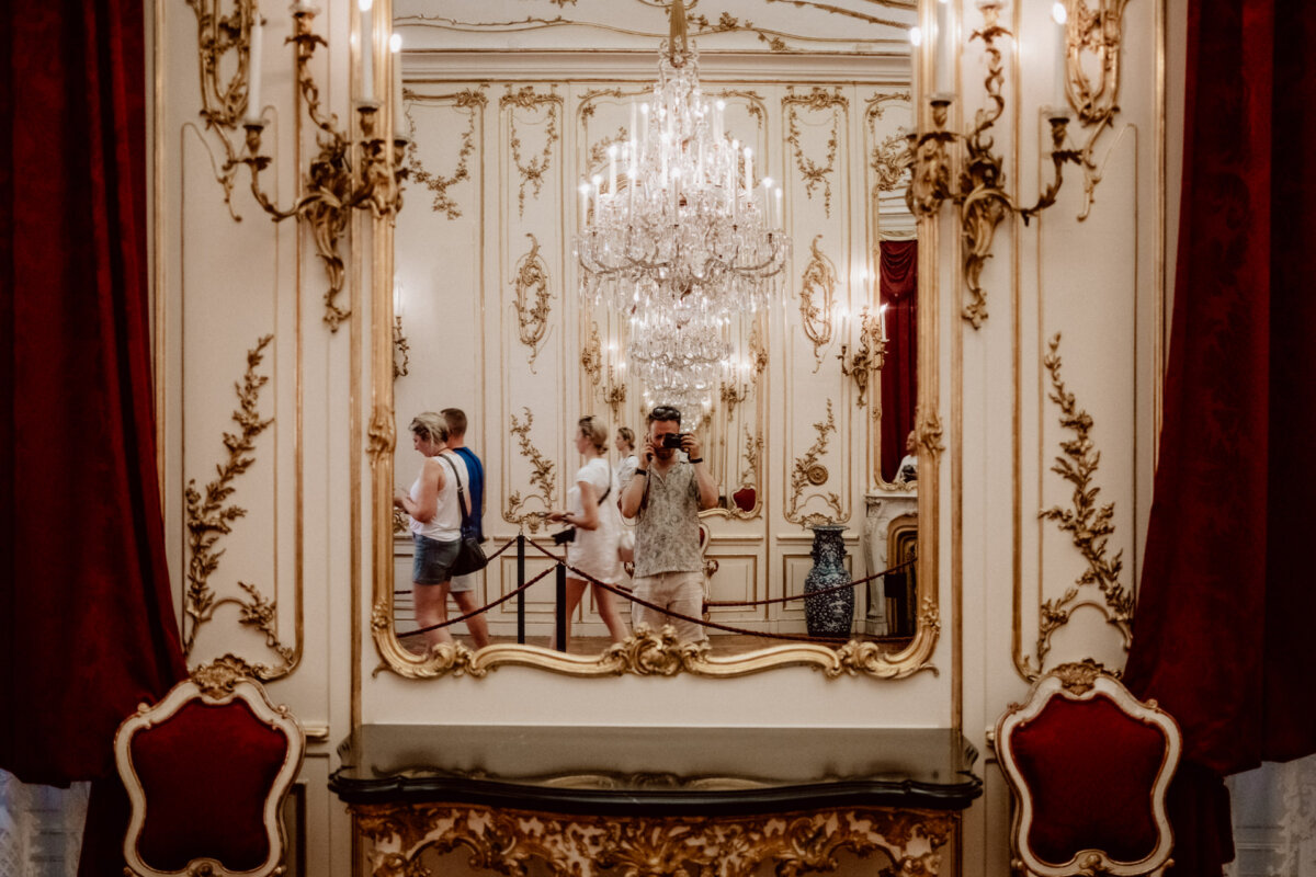 A person takes a picture of a richly decorated room with ornate gold details, red velvet curtains and a large chandelier reflected in a mirror. Other guests are seen walking in the background, admiring the opulent surroundings. 