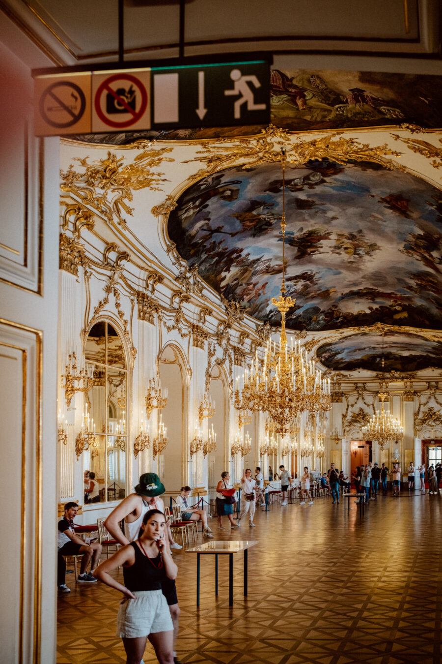 A huge, richly decorated hall with a painted ceiling, large chandeliers and mirrors on the walls. People stand and sit around, admiring the architecture. In the foreground hangs a directional sign with exit symbols and prohibition signs.  