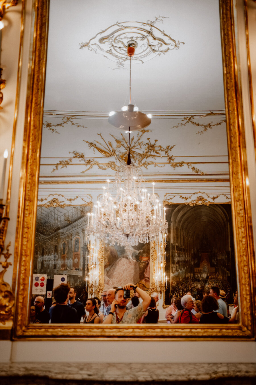 A large, ornate mirror reflects the chandelier and the crowd of people in the lavish room. The room has intricate gold details on the ceiling and walls and a large painting. Visitors seem to be exploring the lavish, historic surroundings.  