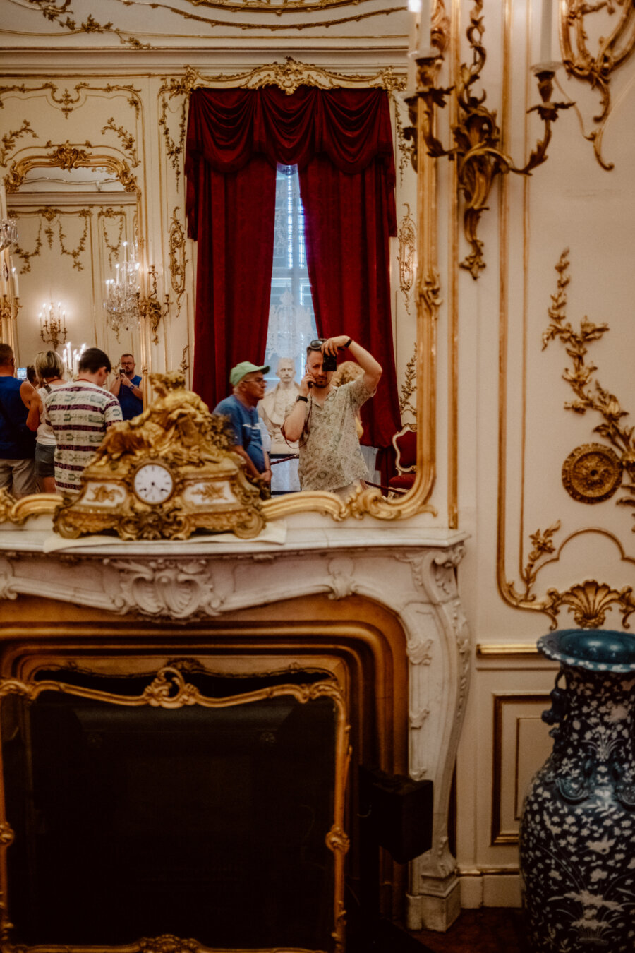 A group of people are looking at an ornate room with gold walls and red curtains. A large mirror above the fireplace reflects the person taking the photo. The room has an antique clock, decorative vases and chandeliers.  