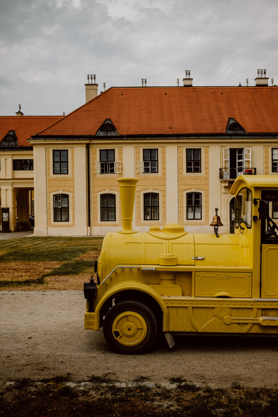 A small yellow streetcar styled as a locomotive stands on a gravel path in front of a large, imposing building with a red tile roof and cream-colored walls. The sky is overcast and the ground has clumps of green grass. 