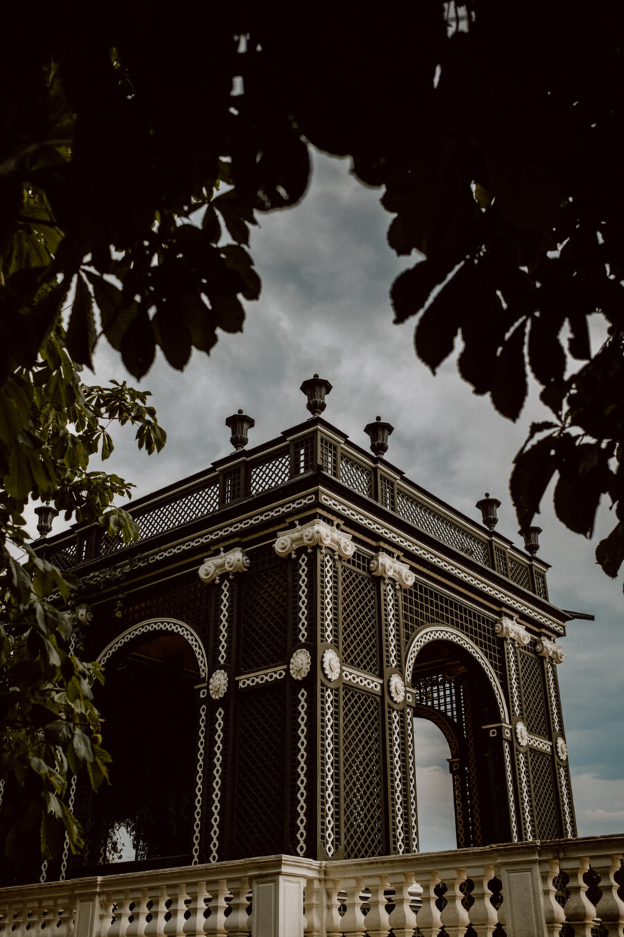 An ornate gazebo with intricate lattice and decorative details stands against a backdrop of an overcast sky. The structure is framed by leafy branches in the foreground, adding a natural accent to the architectural scene. 