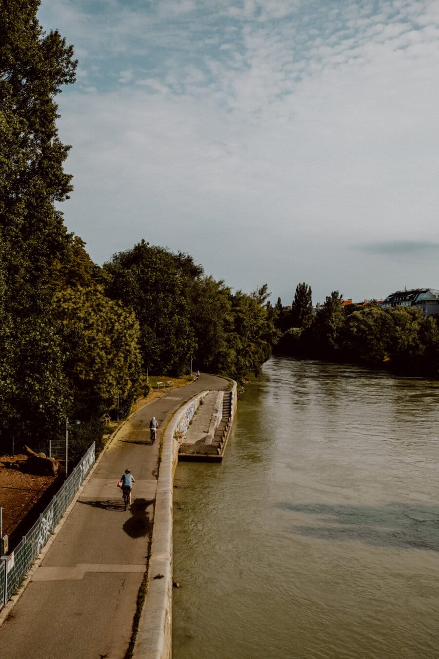 A peaceful path by the river with lush trees on the left and calm water on the right. Two cyclists ride along the path under a partly cloudy sky. The scene exudes a peaceful, natural atmosphere.  