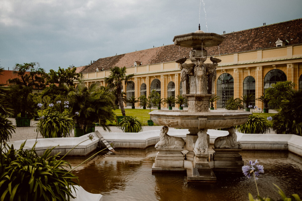 A large, ornate stone fountain with carved figures stands in a peaceful garden surrounded by lush greenery. In the background you can see a long yellow building with arched windows and a red tile roof, with an overcast sky above. 