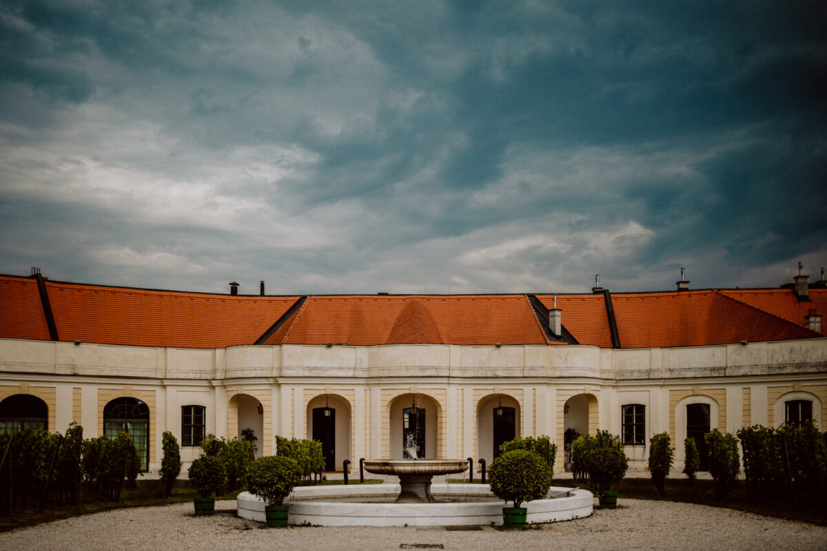 The historic building with its red tile roof and arched facade stands against an overcast sky. In front, a circular stone fountain is surrounded by well-trimmed shrubs in pots, arranged symmetrically in a gravel courtyard. 