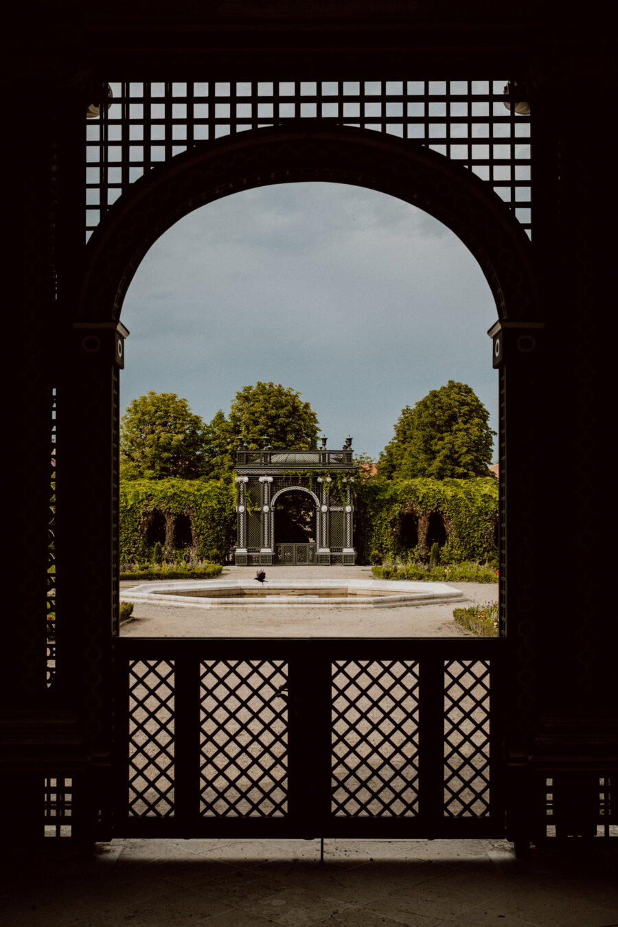 A view through an ornate archway of a garden with a small pavilion in the center, surrounded by lush greenery. The foreground is framed by the elaborate lattice of the arch, leading the eye toward the peaceful garden and pavilion under an overcast sky. 