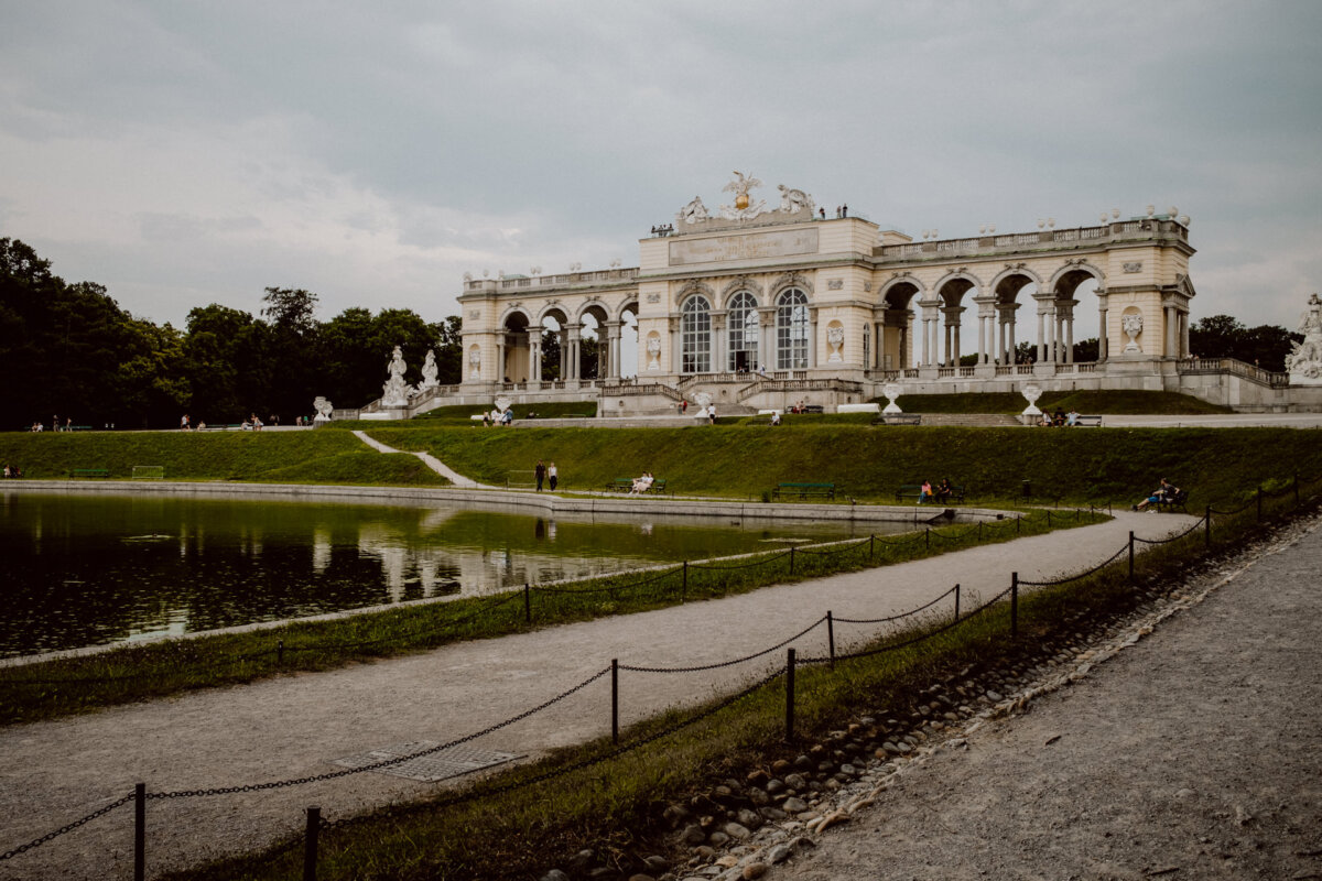 A large neoclassical pavilion with arched windows and columns stands atop a grassy hill. A reflecting pond and paths are in the foreground, while several people walk and sit on benches. The sky is overcast, adding to the scene's peaceful atmosphere.  