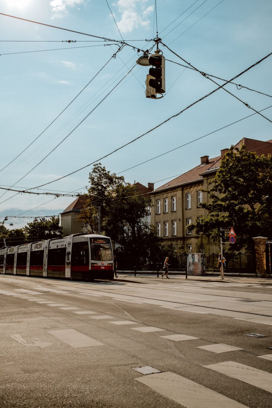 Street scene showing a streetcar traveling along the tracks past a crosswalk. Overhead wires are visible. In the background you can see multi-story buildings among trees under a clear blue sky. Several pedestrians are walking along the sidewalk.   