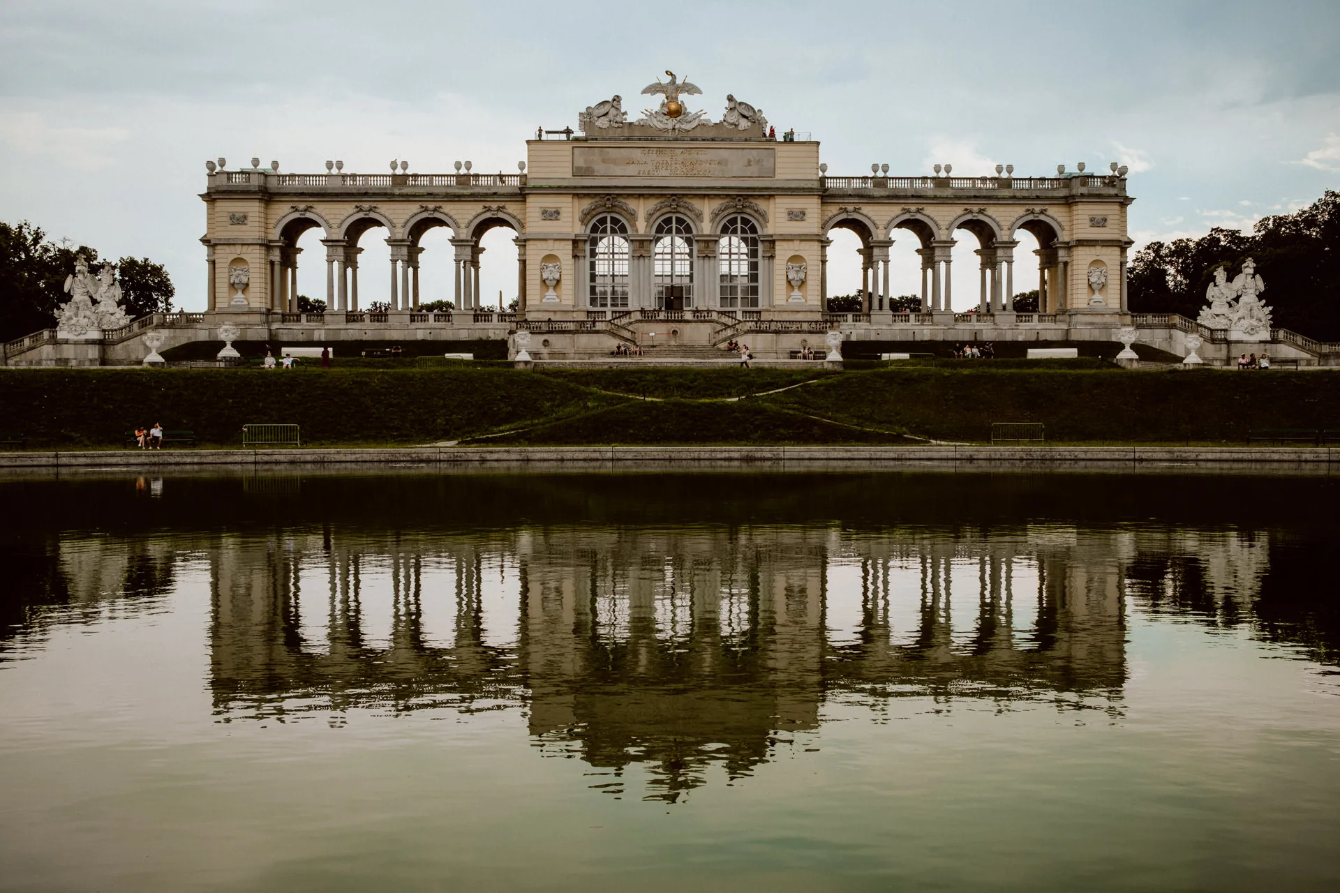 The image shows the Gloriette, a grand arched building with columns and statues, reflected in a calm pond on a cloudy day at Schönbrunn Palace Gardens. People relax on the grass, making it an inspiring scene for fotograf eventowy warszawa.