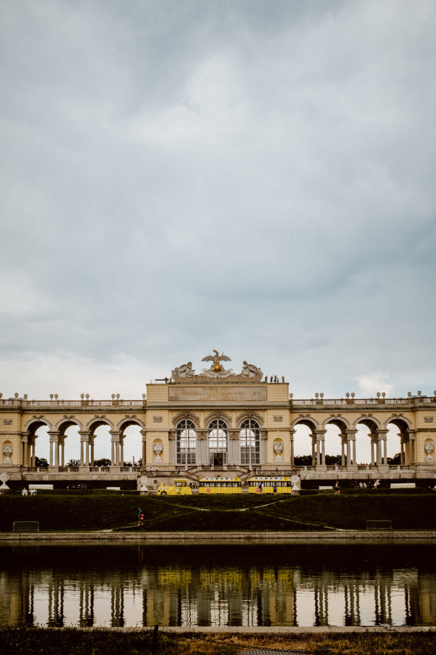 A huge historic building with a series of arches and sculptures stands under an overcast sky, reflected in the calm sheet of water in the foreground. Figures can be seen on top of the structure and walking around it, suggesting a popular tourist attraction. 