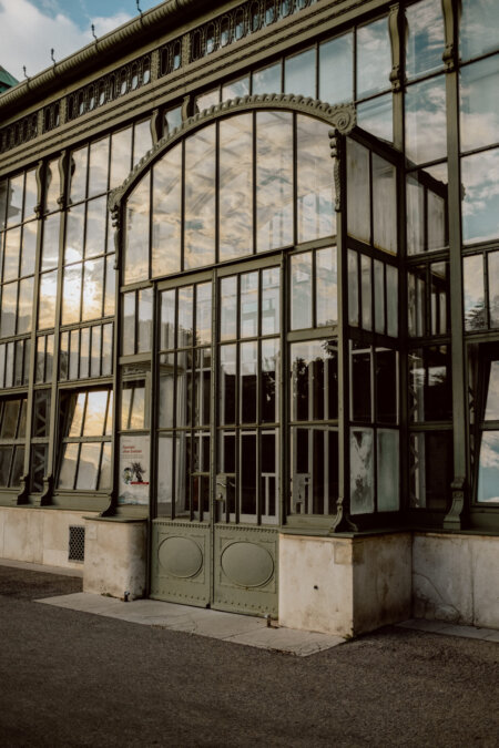 A large, ornate greenhouse with tall glass windows and an intricate metal frame. The entrance has double doors with glass panels. The glass shows the reflection of the evening sky and clouds. The ground in front of the building is paved.   