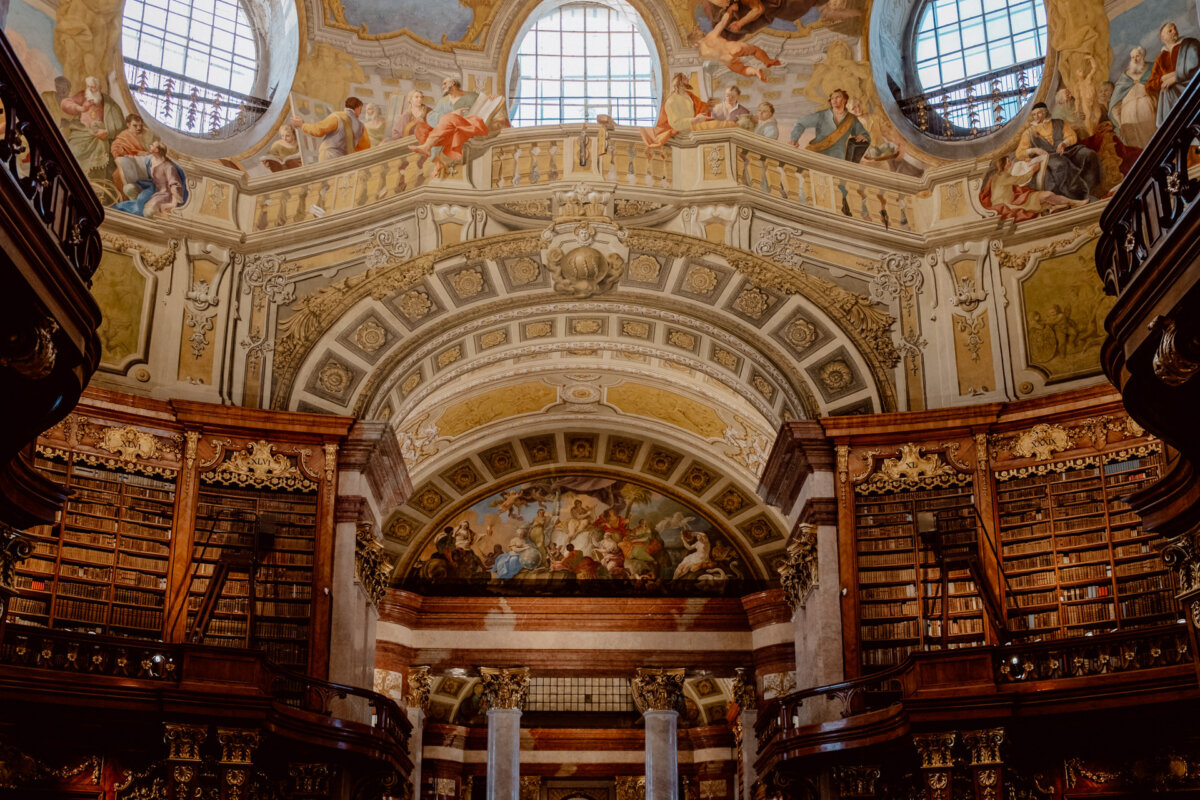 A huge, ornate library interior with tall wooden bookcases filled with books. The ceiling is decorated with intricate architectural details and colorful frescoes. Large arched windows allow natural light to flood the space, accentuating its splendor.  