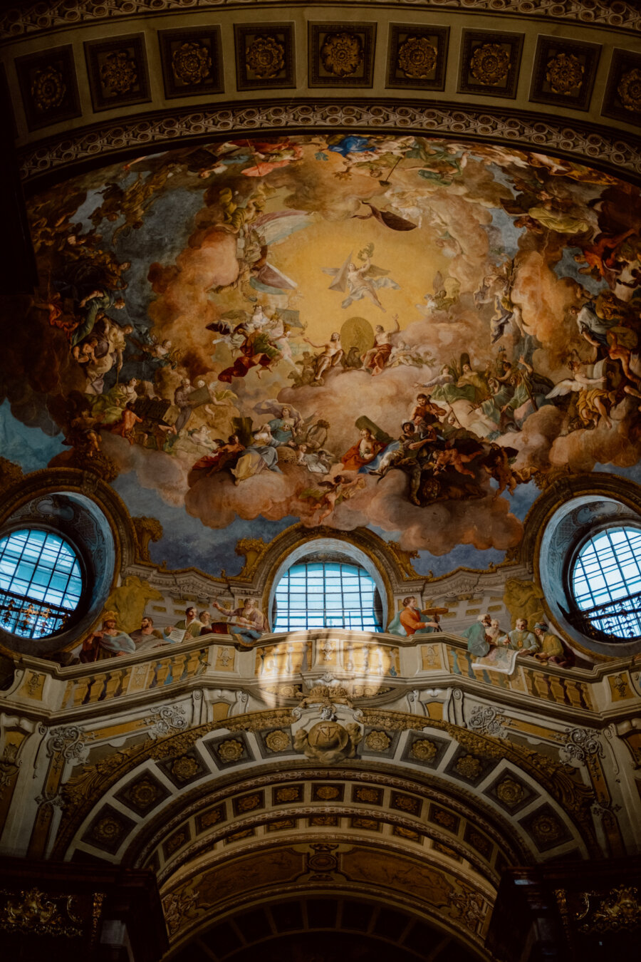 The church's richly painted ceiling displays intricate artwork depicting religious and angelic figures amid clouds and golden hues. Beneath the curved fresco, arched windows and ornate architectural details are illuminated by natural light. 