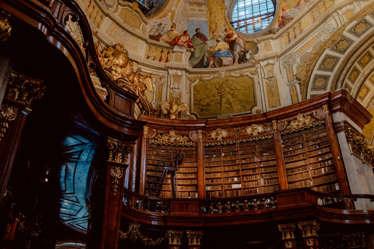 A huge, richly decorated library with an intricately ornamented ceiling featuring classical paintings and sculptures. High wooden shelves are filled with numerous old books, and a ladder leans against one of the shelves. The architectural design is baroque.  