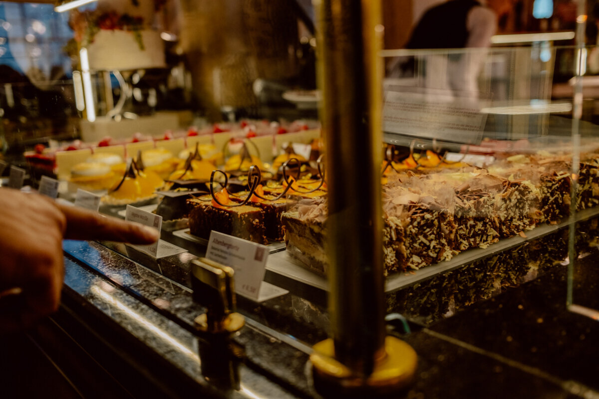 A person points to a selection of different desserts displayed behind a glass display case in a bakery or cafe. The desserts include cakes, tarts and other baked goods, beautifully arranged in rows. The background shows a warm, inviting interior.  