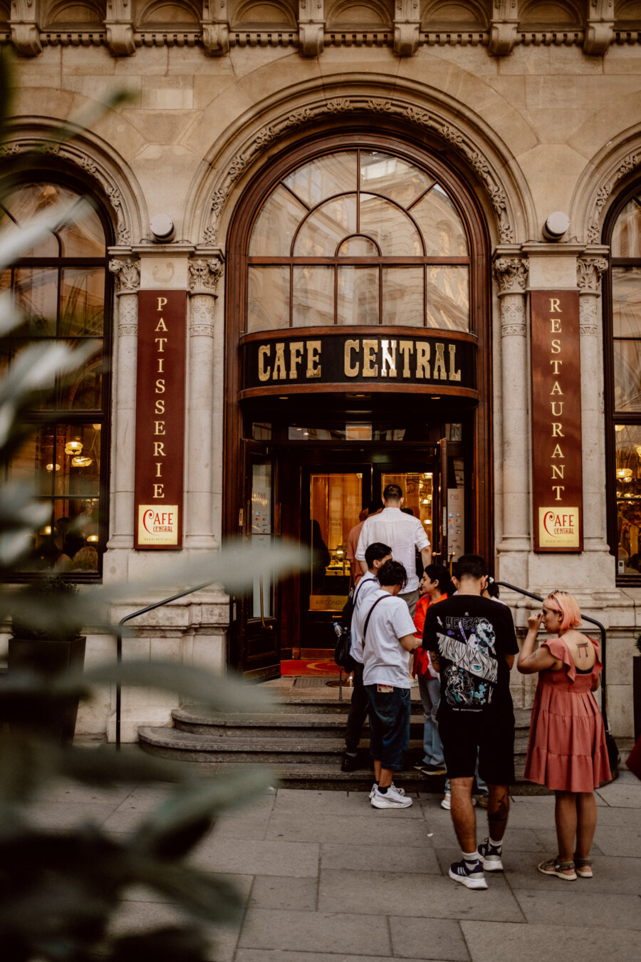 A group of people waiting in line to enter the historic Cafe Central with arched windows and decorative stone features. The cafe has signs reading "Patisserie" and "Restaurant" on either side of the entrance. Lush greenery is visible in the foreground.  