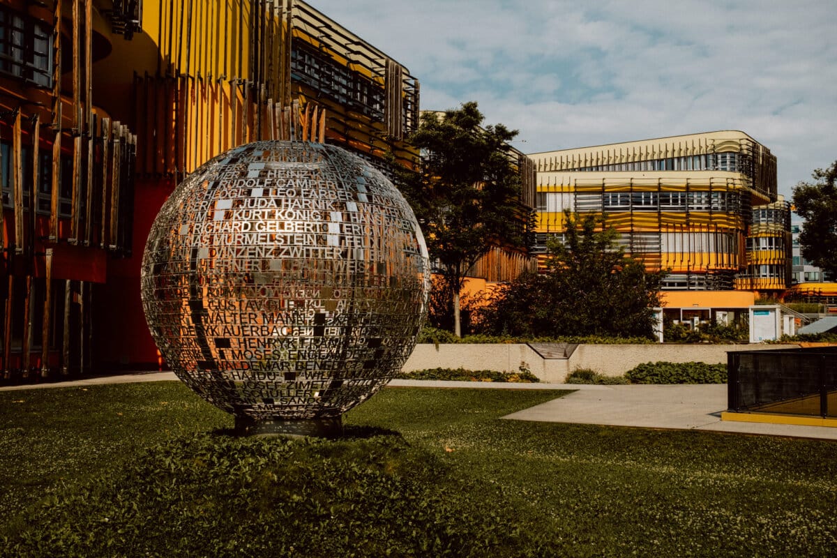 A large spherical sculpture made of metal letters stands on a grassy area with a modern, colorful building in the background. The building has yellow, orange and brown horizontal elements, and trees and shrubs surround the sculpture. 