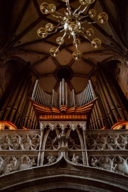 A dramatic upward view of the large church organ, showcasing intricately designed pipes, ornate carvings and a large chandelier with multiple lights hanging from the high arched ceiling above. The architecture shows Gothic influence. 