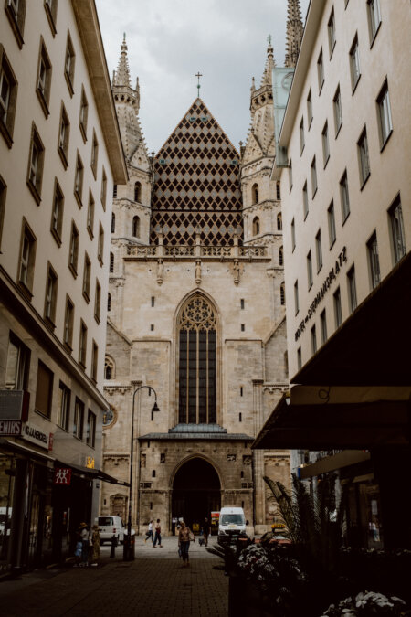 A view of a Gothic-style cathedral with a patterned roof as seen through a narrow street surrounded by tall buildings. Pedestrians and vehicles populate the street, and stores with signs are lined up along the ground level of the buildings. The scene takes place under an overcast sky.  