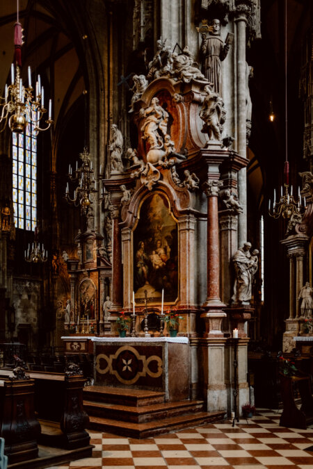 An ornate altar in a historic cathedral, with a large religious painting framed by intricate marble columns and statues. Chandeliers hang from the ceiling and stained glass windows let the light shine through, illuminating the grandeur of the surroundings. 