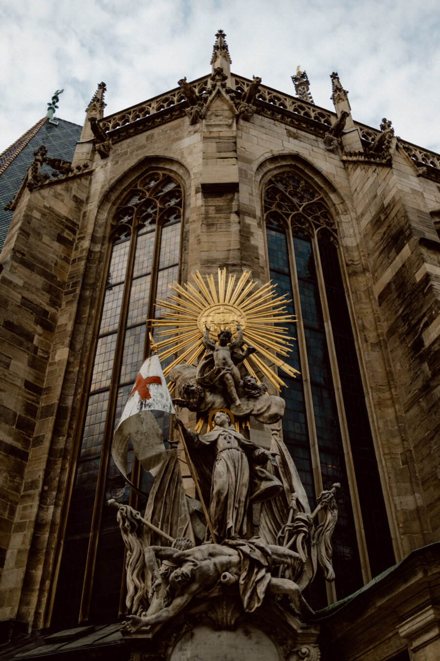 Photo of a detailed religious statue depicting multiple figures in front of the facade of a Gothic-style church. The statue includes a central figure decorated with a golden halo in the shape of a sunbeam and holding a flag with a red cross. The church has tall, arched stained glass windows.  