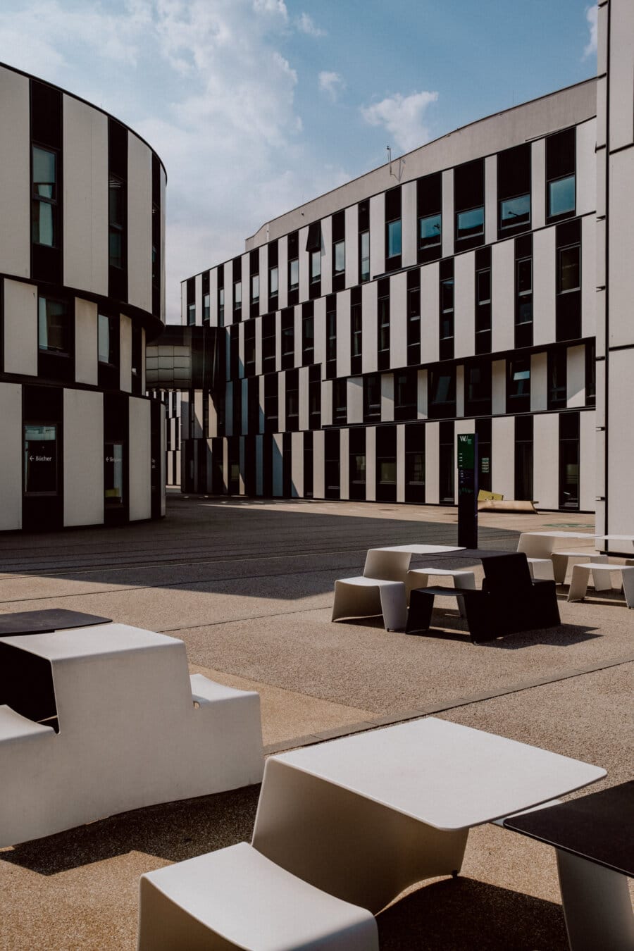 Modern building with white and black geometric patterns, with large windows. The courtyard has uniquely shaped white and black tables and benches. The sky is partly overcast and the atmosphere is peaceful.  