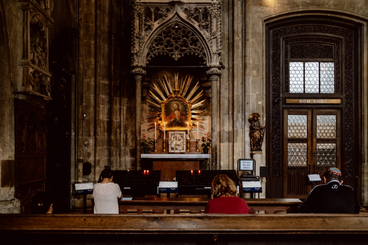 Four people sit in the church facing an illuminated altar with a religious painting in the center. The area has Gothic architectural details, and a wooden door with stained glass panels is on the side. Lighted red candles are visible near the altar.  