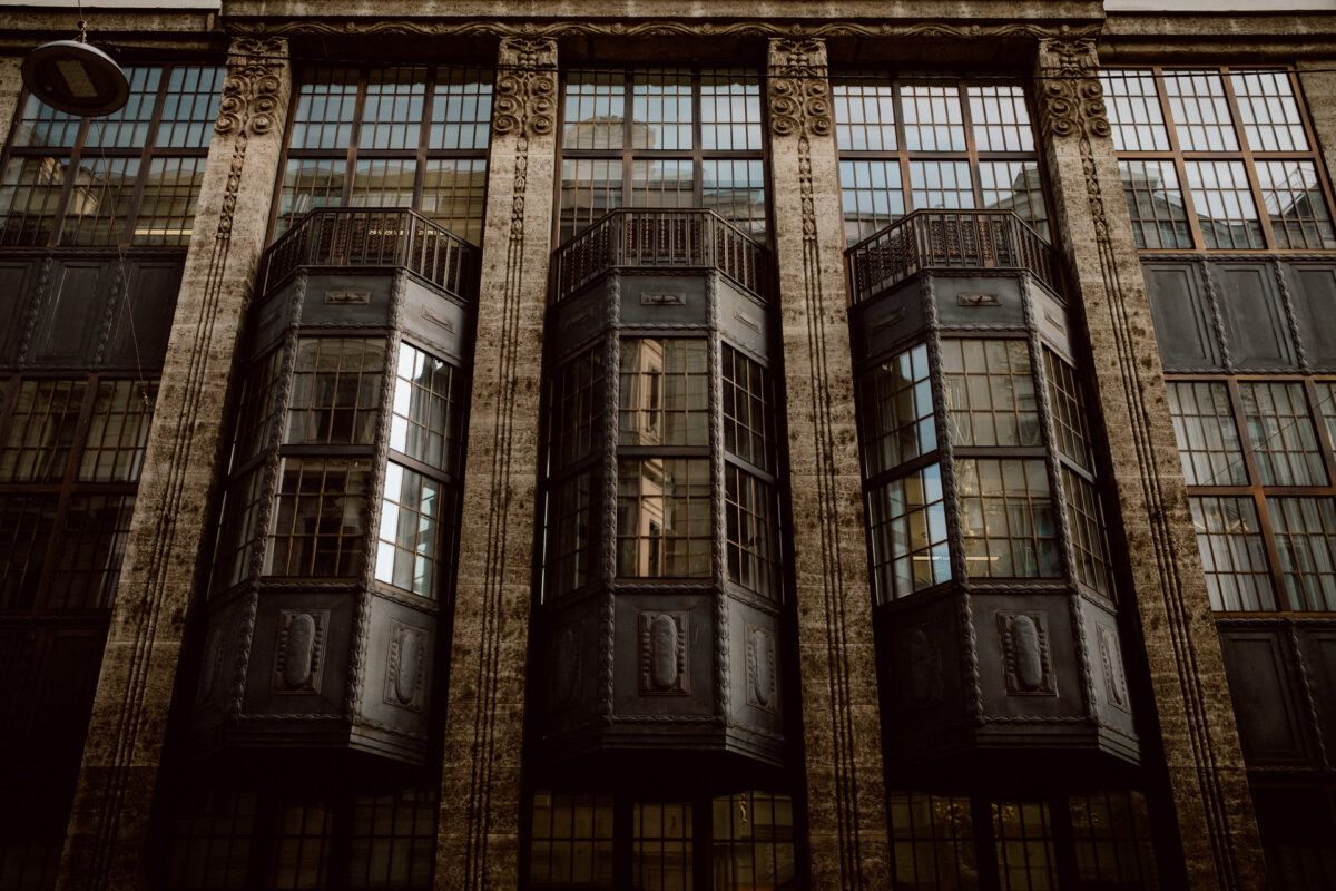 The historic facade of the building features three large, tightly spaced, projecting bay windows with dark metal frames and numerous small panes of glass. The structure is decorated with ornate stone carvings, displaying a historic and industrial aesthetic. 