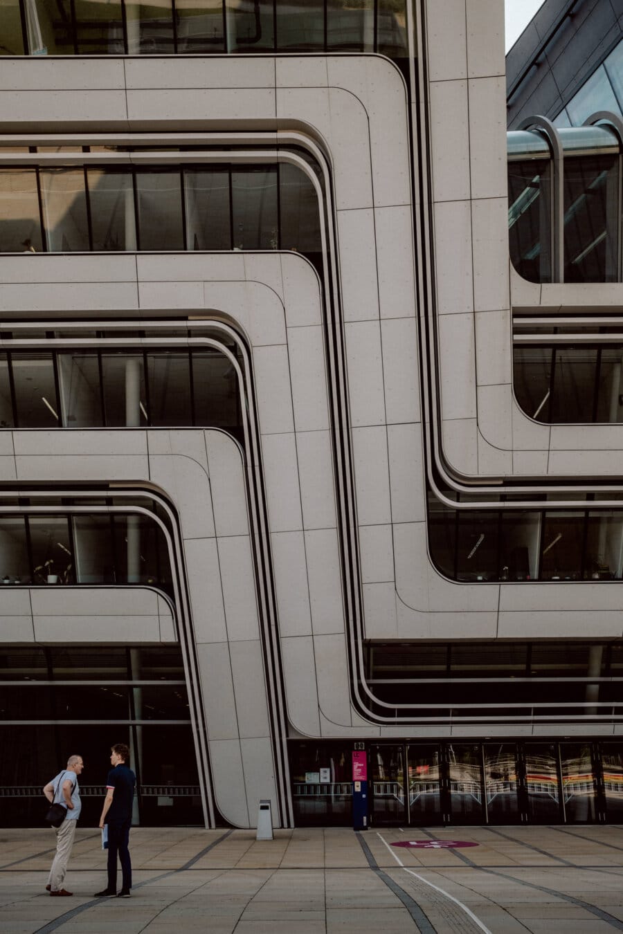 Two people are talking in front of a modern building with a unique curved geometric facade. The building has overlapping white concrete strips with large windows. The ground is paved and the scene appears to be in an urban setting.  