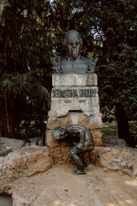 The stone bust of Sebastian Kneipp is placed on a pedestal. Below it is a bronze statue depicting a person leaning over water. On the pedestal is an inscription with Kneipp's name and years of life. The background includes greenery with trees and shrubs.   