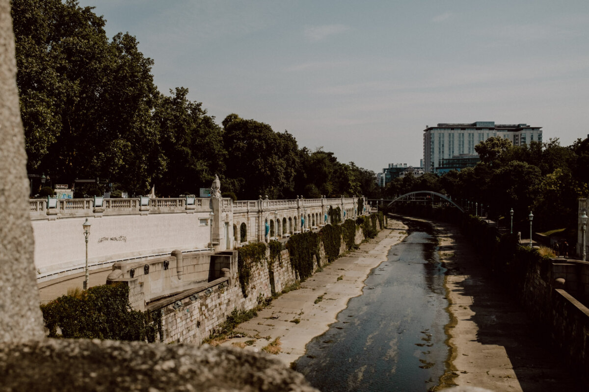 A stone riverbed with rare water flows under an arched bridge. The river is surrounded by trees and an ornamental stone railing on the left. A tall building stands in the background under a partly cloudy sky.  