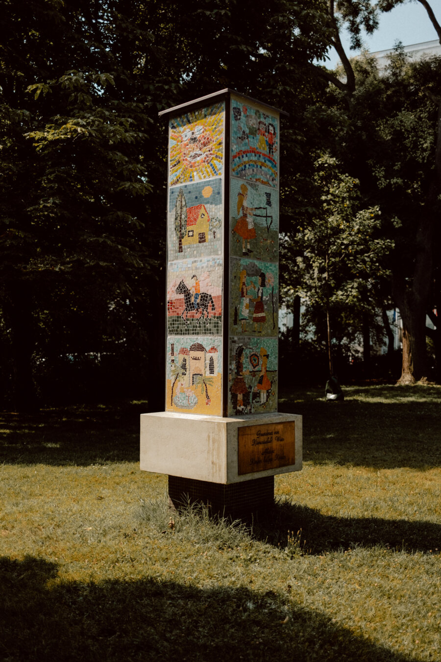 The tall outdoor mosaic column stands on a stone base on a grassy area. The column features colorful, detailed panels depicting various scenes, including houses, people and landscapes. Sunlight seeps through the trees in the background.  