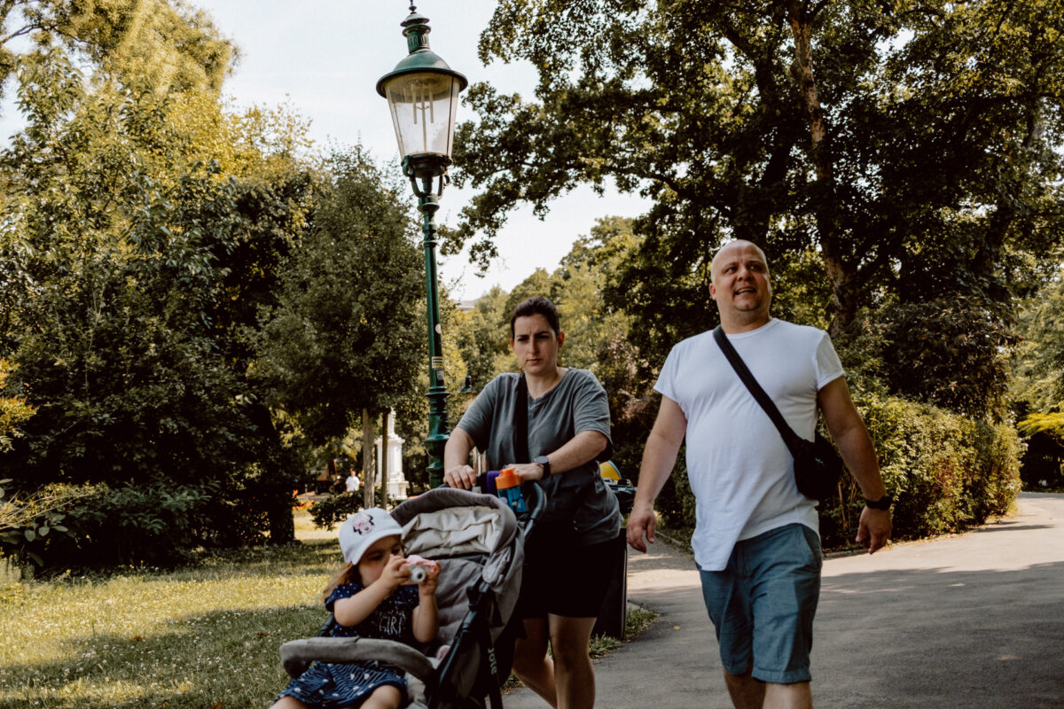 A man and a woman are walking along an alley in a park, pushing a stroller with a small child. The woman is on the left with the stroller, and the man is walking beside them on the right, both dressed in everyday summer clothes. A lamppost can be seen in the background.  