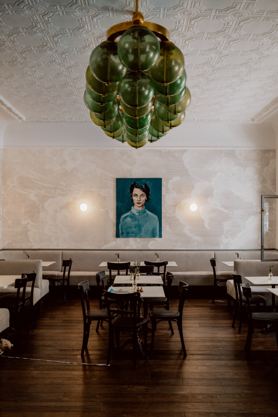 An elegant, modern dining room with dark wooden tables and chairs, plush white booths and a large green chandelier. A blue portrait of a woman sits at the center of a patterned white wall. The space has a warm, welcoming atmosphere with modern decor.  