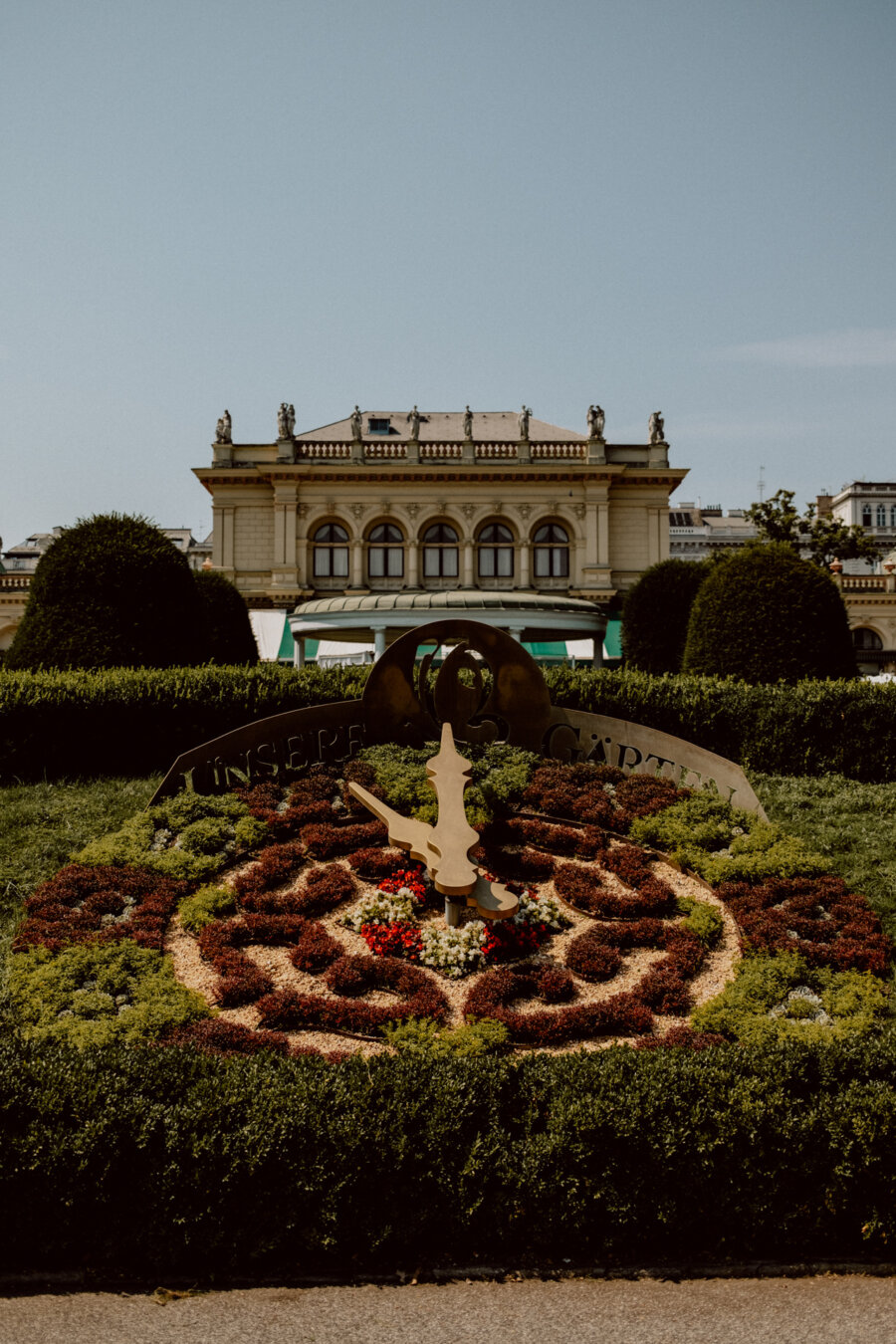 A large decorative garden clock with colorful plants and flowers in front of a historic classical building decorated with statues and architectural details. The clock has a metal dial and the word "UNSERE" is partially visible. 