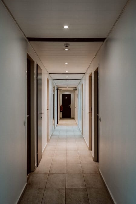 A narrow, well-lit corridor with white walls and a tiled floor. Several closed doors on either side of the hallway and a dark door with the number "20" at the end. Overhead lighting evenly illuminates the passage.  