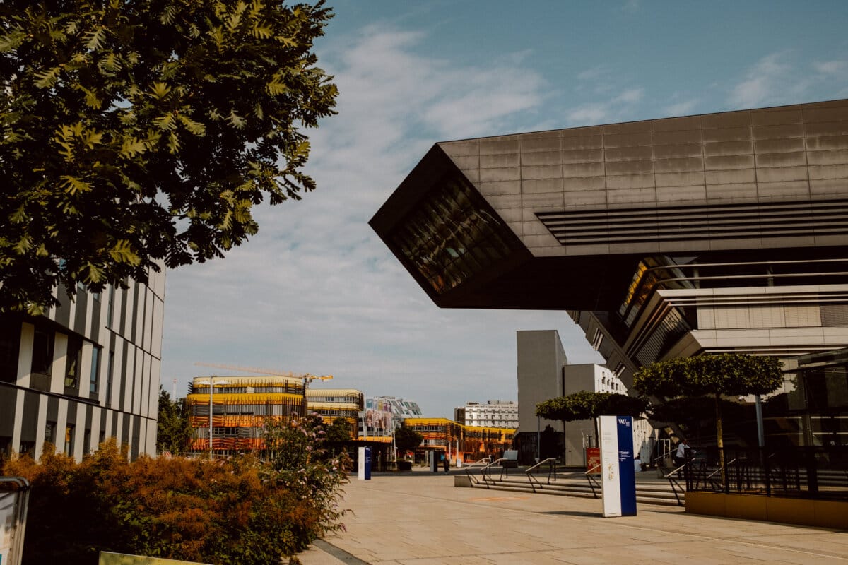 An open square with modern buildings with unique geometric patterns and reflective surfaces, surrounded by trees and shrubs. In the background are more contemporary buildings with colorful accents under a blue sky with scattered clouds. 