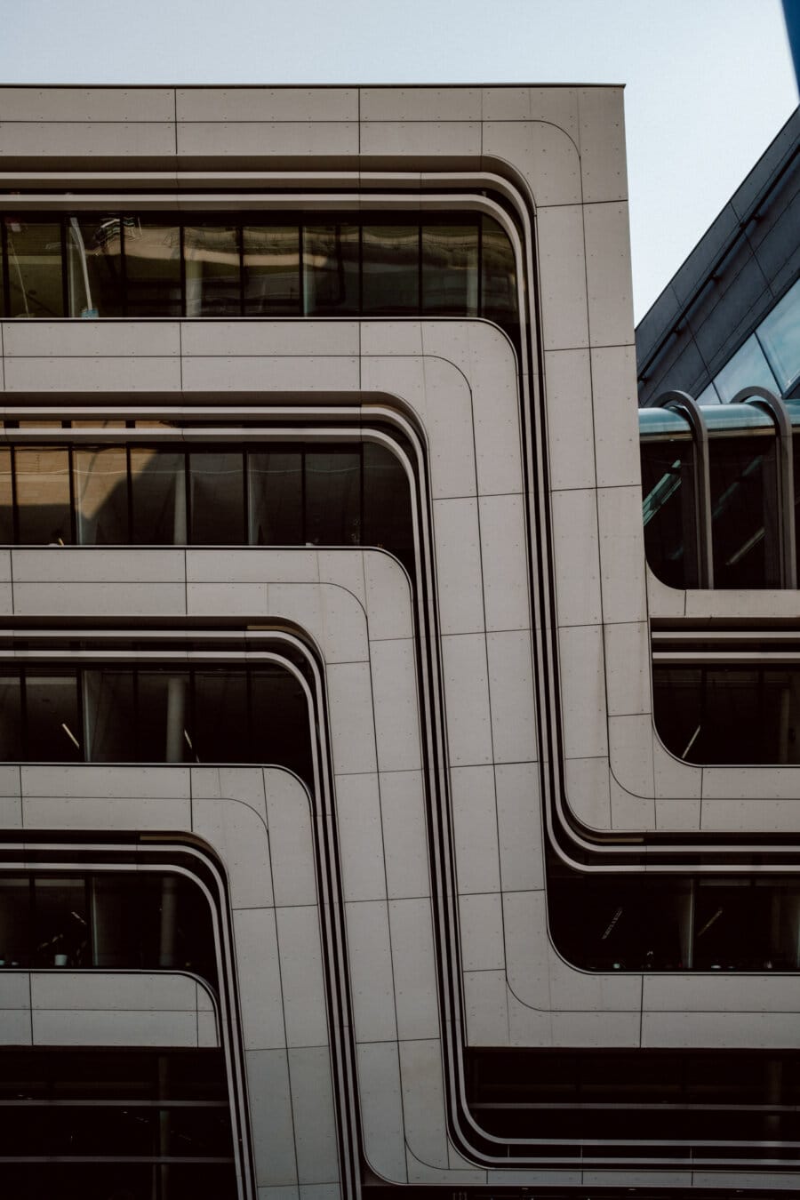 A modern building facade with a unique design consisting of multi-layered, undulating and angular glass and concrete sections. The building presents long horizontal lines that create a visually striking geometric pattern under a clear sky. 