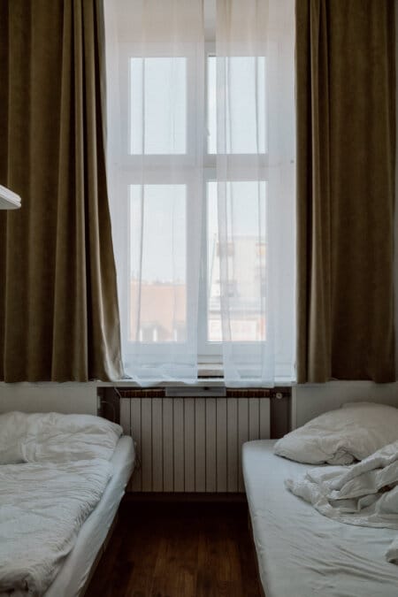 The dimly lit room has two unmade beds parallel to each other, separated by a radiator under a large window. The window is covered with thin white curtains and flanked by heavier beige curtains. Wooden flooring adds a warm touch to the room.  